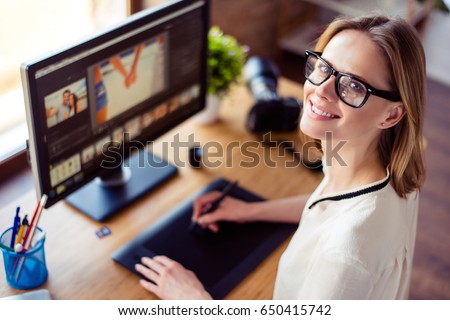 Top view of intelligent blond young woman working with computer and graphic tablet, stylus, smiling. She is a successful self employed retoucher and photograph