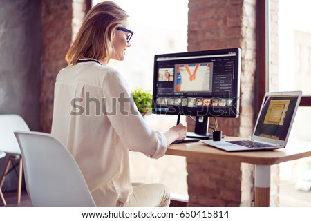 Smilimg blond girl is working at home on computer, using her laptop also. She is a graphic designer. Retouch is her hobby, she is self employed freelancer Royalty-Free Stock Photo #650415814