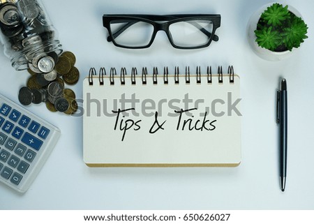 Top view of calculator, coins, jar, eye glasses, plant, pen and open notebook written with TIPS AND TRICKS on white background. Business concept.