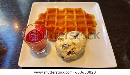 Fresh Belgium waffle with vanilla ice cream with chocolate bits and raspberry jam in a glass on a square plate.