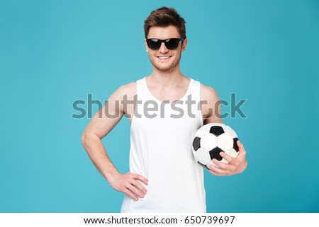 Picture of young cheerful man standing over blue isolated background holding foot ball. Looking at camera.