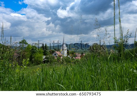small szekler village with white steeple among grren hills, cloudy sky in the spring / East European villages - Transylvania region