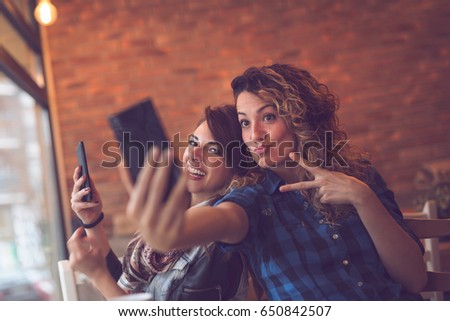 Two friends drinking coffee in a cafe, taking selfies with a smart phone and having fun making funny faces. Focus on the girl on the right