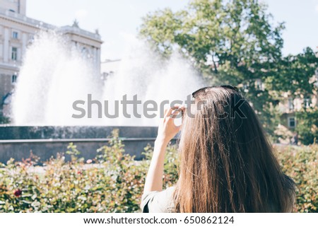 Young woman with long brown hair taking pictures of the fountain in the park