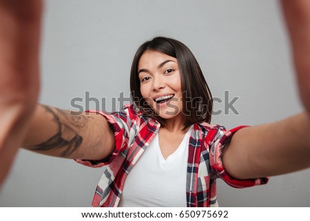 Picture of young cheerful woman make selfie over grey wall. Looking at camera.
