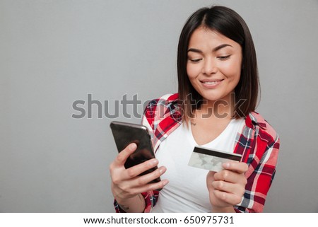 Picture of young happy woman holding credit card and using mobile phone over grey wall. Looking aside.