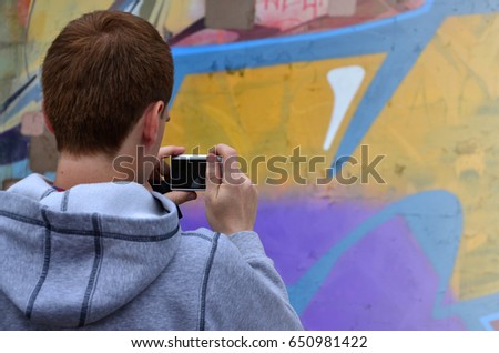 A young graffiti artist photographs his completed picture on the wall. The guy uses modern technology to capture a colorful abstract graffiti drawing. Focus on the photographing device