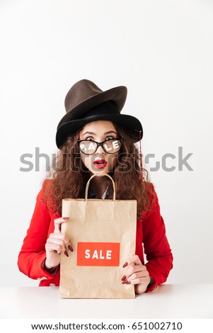Photo of shocked young caucasian lady sitting isolated over white background holding shopping bag wearing a lot of hats by sale. Looking at camera.
