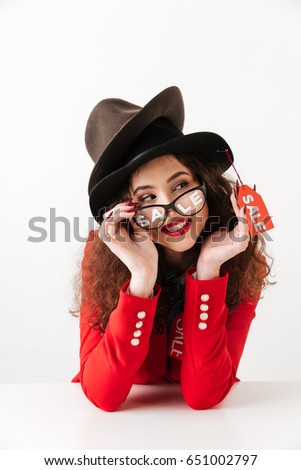 Picture of smiling young caucasian lady sitting isolated over white background wearing a lot of hats by sale. Looking aside.