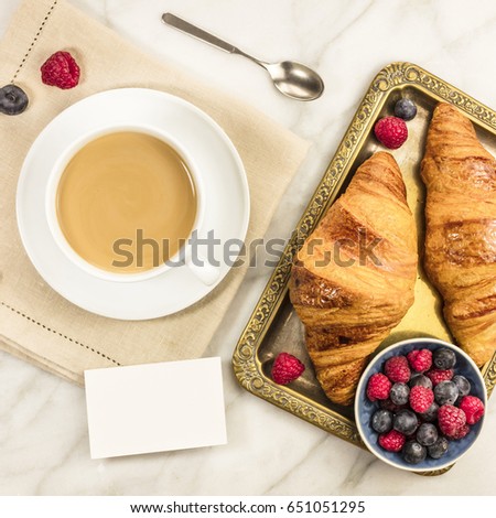 A square photo of a cup of coffee with fresh raspberries, blueberries, and croissants, with a blank business card with a place for text