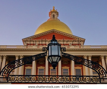 Closeup at entrance to Massachusetts State House in Boston includes gold dome, some of the building's facade and the front gate in the foreground that says in gold letters: Massachusetts State House.
