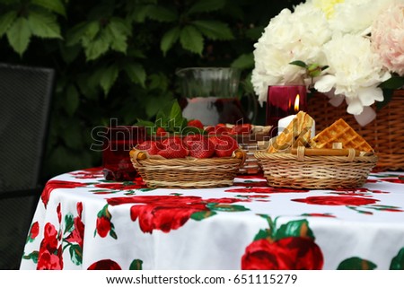 White peonies in a wicker basket. Ripe strawberries and strawberry drink. White tablecloth with red roses. Romantic dinner with candles.
