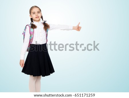 Beautiful little blond schoolgirl, with long neatly braided pigtails. In a white blouse and a long dark skirt.She shows her thumb. The gesture is okay.On the pale blue background.