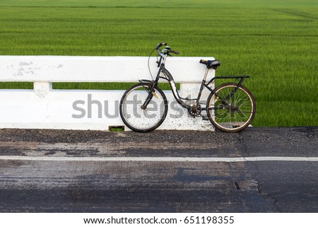 An old bicycle with a white concrete bridge on a paved road with green rice fields as a backdrop.