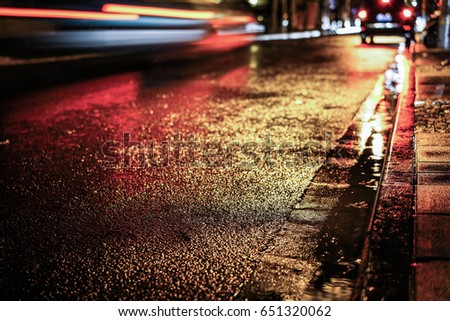 Rainstorm night in the big city, light from the shop windows reflected on the road on which cars travel. View from the level of asphalt.