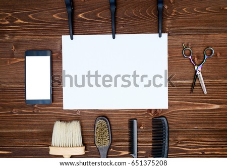Hairdresser tools on wooden background. Blank card with barber tools flat lay. Top view on wooden table with scissors, comb and brush with empty white paper and phone, copy space