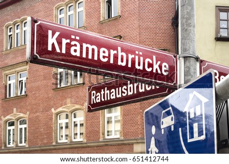 Germany, Thuringia, Erfurt: Close up of deep red street sign (Kraemerbruecke - Merchants' bridge) in the city center of the famous German town with red brick house facades in the background - travel