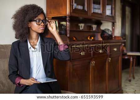 Serious black businesswoman sitting in the room with the tablet in her hands. Woman wearing suit and glasses.