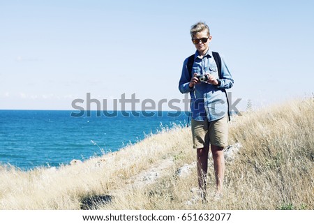 Summer travel concept. Happy young traveler hipster with backpack holding photo camera and take a picture at sunshine shore near beach