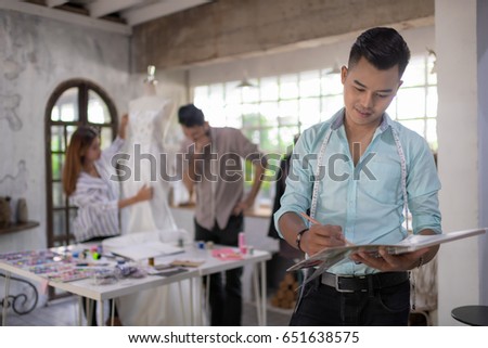 Smiling Wedding dressmaker designer using digital tablet with blur team working on mannequin in wedding dress and equipment background.
