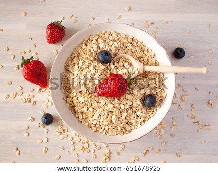 oat groats in white ceramic bowl with wooden spoon, strawberries and blueberries and oats scattered on the table