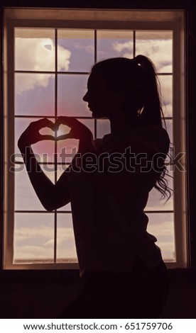 A profile of girl which is standing by window and making heart with her hands. Romantic mood. Sunset. Silhouette. Backlight.
