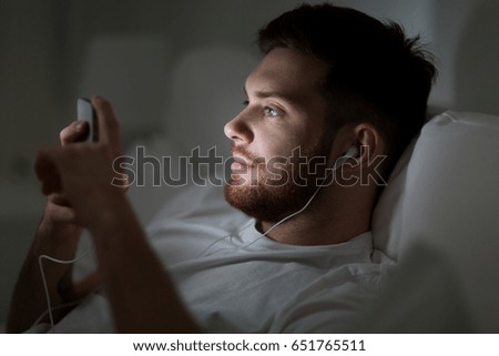 technology, internet, communication and people concept - happy smiling young man with smartphone and earphones listening to music in bed at night