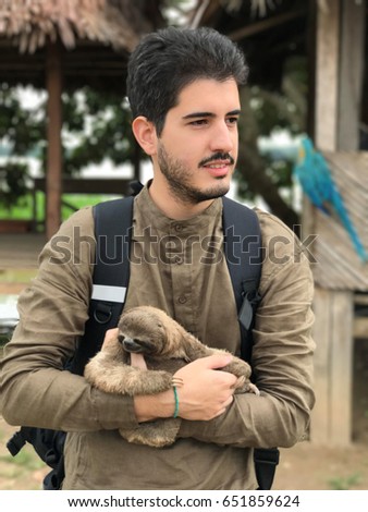 Adventure travel concept. Young white man holding a sloth at the Amazon jungle, blurred shacks at background