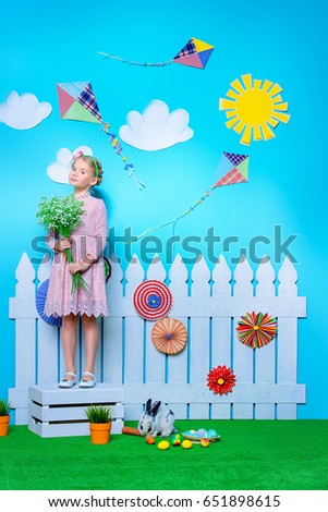 Cute happy little girl standing with Easter Bunny in spring decorations. Easter Bunny and painted eggs. Kid's fashion. 
