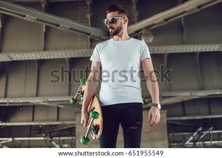 Stylish man in sunglasses and with a beard stands on the street with a long board. T-shirt mock up. Royalty-Free Stock Photo #651955549