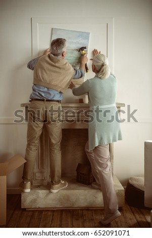 rear view of senior husband and wife hanging picture on wall over the fireplace