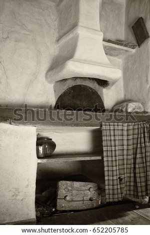 An old house. Fragment of a wood stove with a chimney and a fireplace. In the frame there is a shelf, a curtain, dishes, a pot, a stool. Black and white image.