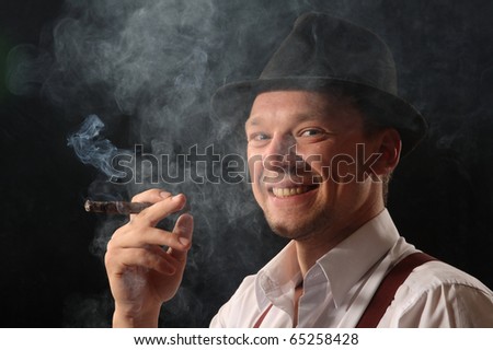 Portrait of the young man against a dark background in the black hat, smoking the big cigar.