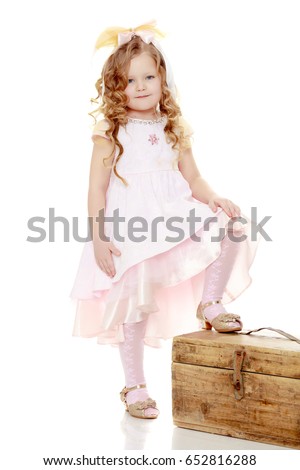 Beautiful, chubby little girl with long, blond,curly hair.Dressed in a pink dress and a bow in her hair.The girl put his foot on an old suitcase.Isolated on white background.