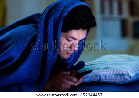 Social media addict men on bed not sleep because play smart phone, covering his head with a blue blanket, room background