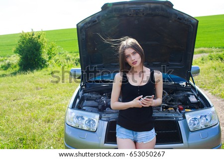 Closeup portrait, young woman in pink tanktop having trouble with her broken car, opening hood and texting for help on cell phone, isolated green trees and shrubs outside background
