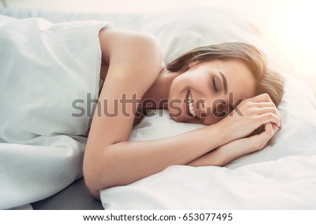 Good morning! Attractive young woman is lying on bed with her hands near face and smiling with closed eyes