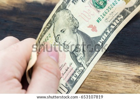 a picture of a Caucasian hand holding a ten USA dollar note on a wooden background.  This image can be used to represent payment.