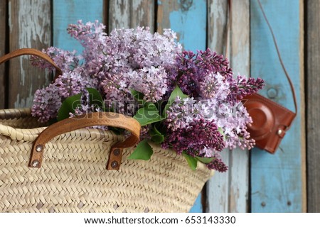 Bouquet, bunch of lilac in vintage authentic wicker farm basket , french style, leather case for camera on blue and grey weathered, aged wooden background, floral composition, daylight, original photo