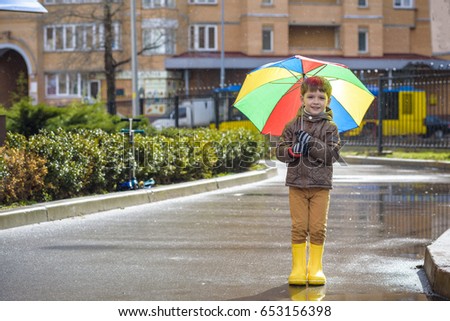Little boy playing in rainy summer park. Child with colorful rainbow umbrella, waterproof coat and boots jumping in puddle and mud in the rain. Kid walking in autumn shower Outdoor fun by any weather.