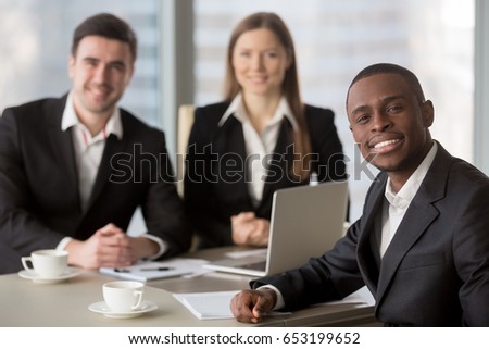 Attractive afro american cheerful businessman looking at camera sitting at desk, young african manager leading meeting for subordinates at background, business consulting, mixed-race team leadership 
