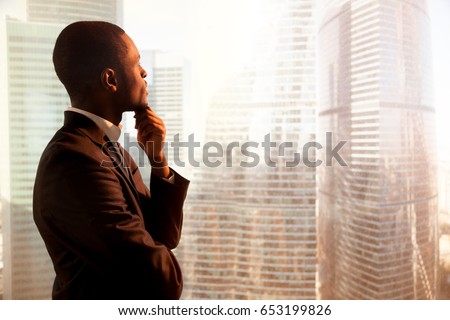 Young african-american thoughtful businessman wearing suit holding hand on chin, looking out of big office window at sunset city building lost in thoughts, planning future project. Side view copyspace Royalty-Free Stock Photo #653199826