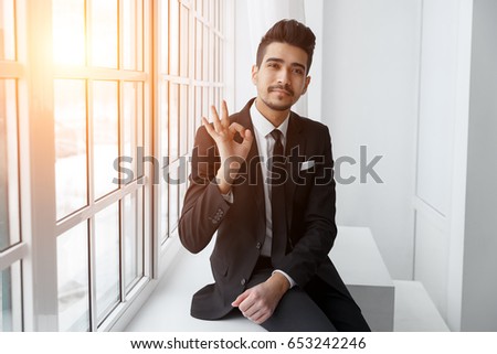 Happy young businessman showing ok sign over office background, sunlight. Business success concept.