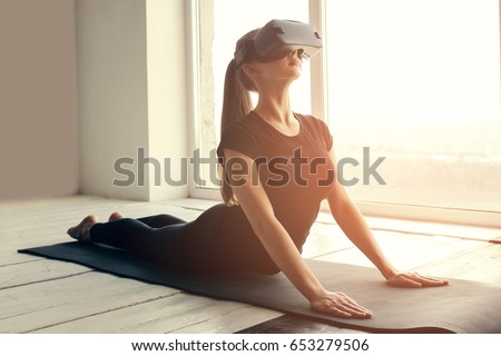 A young beautiful girl in virtual reality glasses makes yoga and aerobics remotely. Future technology concept. Modern imaging technology. Classes in single sports remotely. Royalty-Free Stock Photo #653279506