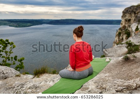 Rear view of young woman sitting in lotus position on rock over the river.