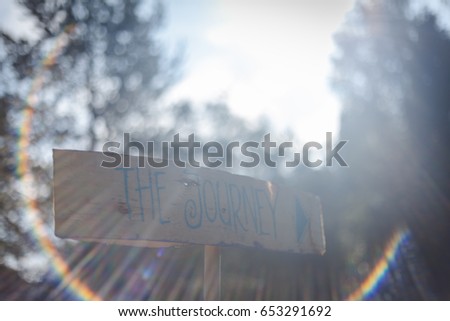 Wooden signpost lit by sun light surrounded by sun flare with the words the journey and an arrow pointing right handwritten on it