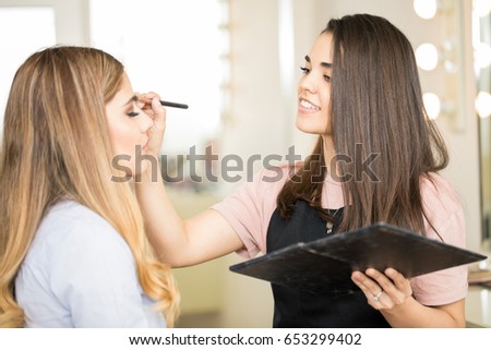 Beautiful young makeup artist applying some eyeshadow on a female customer in her beauty salon