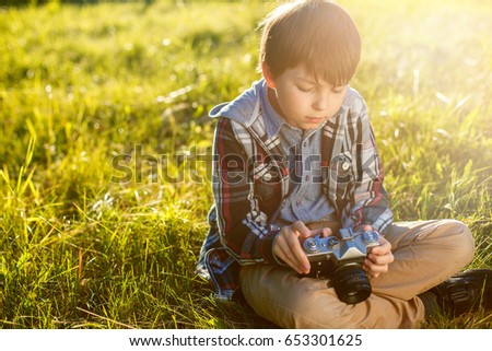 Little boy sitting on green grass in the park and holding camera in hands. Child taking picture using vintage film camera.