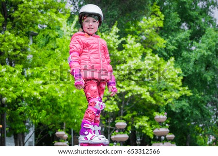 A sporty little girl in a helmet and protective gear stands on roller skates in a city park. Beautiful baby in a pink jacket and rollers is on the background of green trees