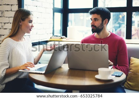 Beautiful female freelancer explaining task to young bearded it developer about creating internet website using modern laptops for work.Students sitting with computers in cafe and discussing ideas 